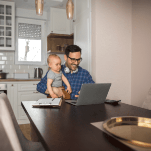 Man working on a laptop in a kitchen whilst holding a toddler